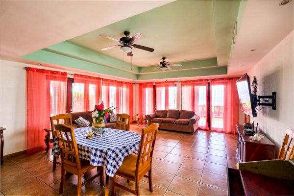 A spacious living and dining area with a checkered table, chairs, a sofa, ceiling fans, a wall-mounted TV, and large windows with orange curtains ending the sentence.