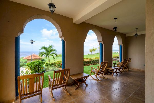 The image shows a covered patio with wooden chairs facing a beautiful outdoor landscape featuring arches, palm trees, and an ocean view in the distance.