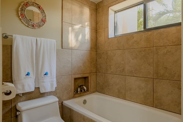 This image shows a bathroom with a bathtub, toilet, towel rack, and window. The walls and floor are tiled, and there are two white towels hanging.