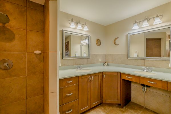 The image shows a modern bathroom with a tiled shower, two sinks with mirrors above, and wooden cabinetry below, all under bright lighting.