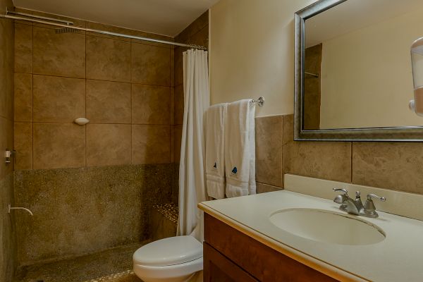 A bathroom with a shower, toilet, wooden vanity with sink, mirror, light fixture, and towels on the rack, all with a beige tile floor.