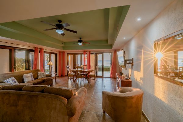 This image shows a cozy living and dining area with comfortable seating, a table, ceiling fans, and a large window with a view of the outdoors.
