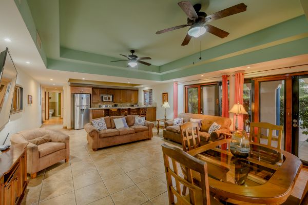 A cozy living and dining area with wooden furniture, beige couches, green ceiling, ceiling fans, and an open kitchen in the background.