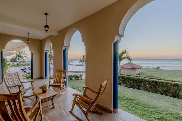 A porch with arched openings, wooden rocking chairs, and a view of a garden leading to the beach and ocean at sunset ending the sentence.