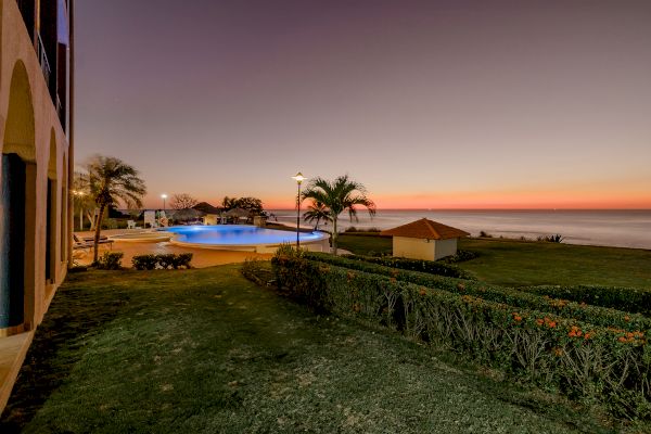 A serene coastal scene at sunset with a pool, palm trees, a small pavilion, and well-manicured lawns overlooking the calm ocean.