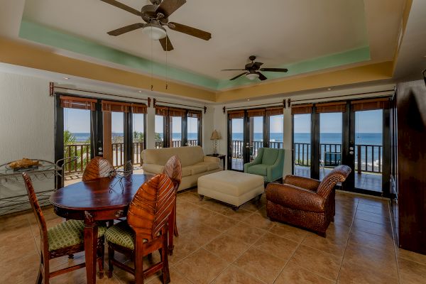 A spacious living room with multiple windows offering an ocean view, comfortable seating, ceiling fans, a dining table, and tiled flooring.