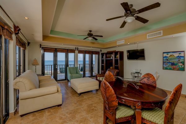 A spacious living room with beige furnishings, a dining table with chairs, large windows, ceiling fans, a wall-mounted TV, and an ocean view ends the sentence.