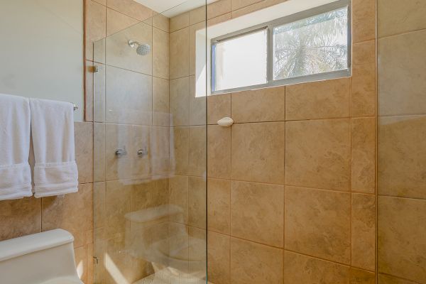 This image shows a modern bathroom with a toilet, glass-walled shower, towel rack, and a window allowing natural light in, all tiled in beige.