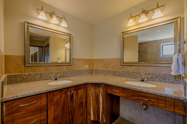 The image shows a modern bathroom with dual sinks, granite countertops, wooden cabinets, and two mirrors, each with three-light fixtures above them.