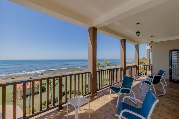 A balcony overlooking a coastal view with several lounge chairs and a small table. The ocean waves can be seen in the distance under a clear sky.