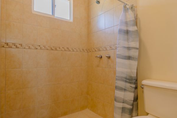 The image shows a bathroom with a toilet, tiled shower area, small window, and a shower curtain. The walls and floors are beige-colored.