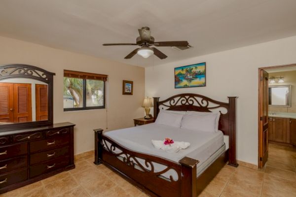 A bedroom features a large wooden bed, dresser, nightstand, ceiling fan, window, and a door leading to a bathroom. Artwork decorates the wall.