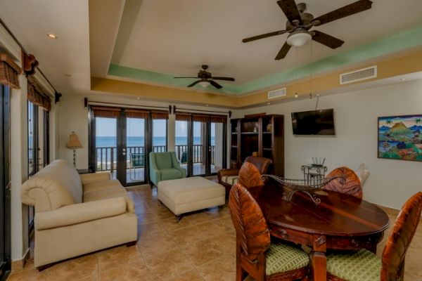 A spacious living room features ceiling fans, large windows with ocean views, two sofas, a round dining table, and a wall-mounted TV ending the sentence.