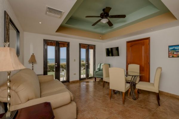 A cozy living room with beige sofas, a wooden table, dining set, ceiling fan, TV, and door. Large windows offer a view of the ocean.