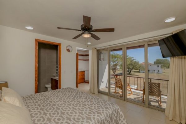 A bedroom with a ceiling fan, patterned bedspread, wall-mounted TV, en-suite bathroom, large sliding glass doors, and a balcony with outdoor seating.