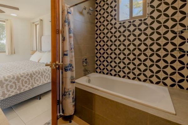 The image shows a bathroom with a bathtub and patterned tiles on the wall. An open door reveals an adjoining bedroom with a bed and matching linens.