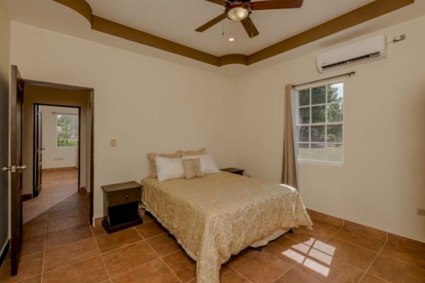 A bedroom with a ceiling fan, air conditioner, bed with beige bedding, and two bedside tables. An open door shows a hallway and another room.