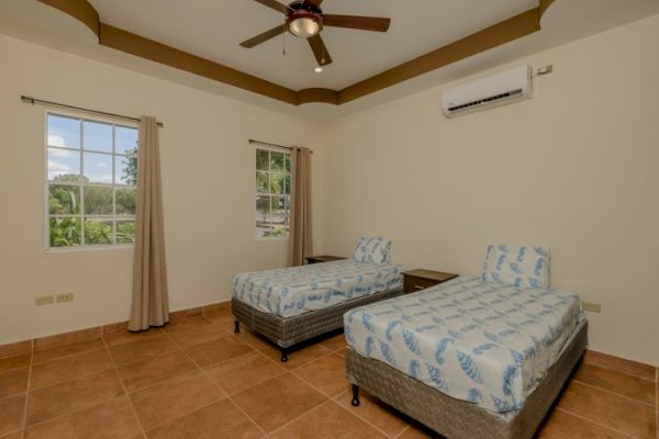 A room with two twin beds, patterned bedding, ceiling fan, air conditioner, and two windows with curtains. Light tiled floor with wooden bed frames.